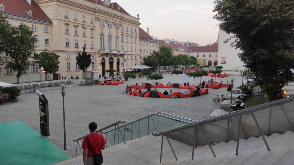 In this picture you can see people seating in a big city. They are sitting on modern benches, they were placed between old and new buildings. In this square the floor is paved, but trees have been planted all around. 

The interpretation is: in a European city that has a long history, we can witness a constant process of renovation that leads to a fusion of old and new elements, an effort to make public spaces accessible to everyone through design. 

On the other side, this pictures shows the “want it all” attitude of people that desire nature, culture, entertainment, noise and peace and above all choices all at once: leaving in cities.